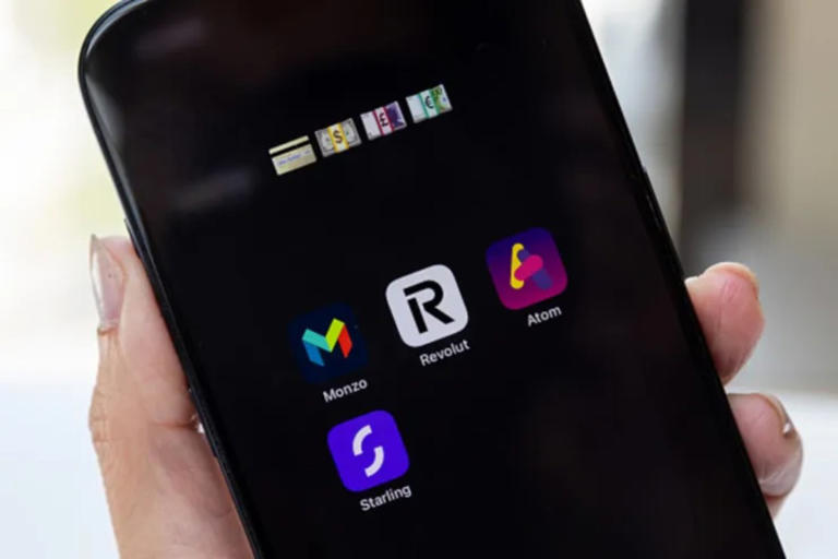 A hand holding a smartphone displays app icons for Monzo, Revolut, Atom, and Starling on its screen.
